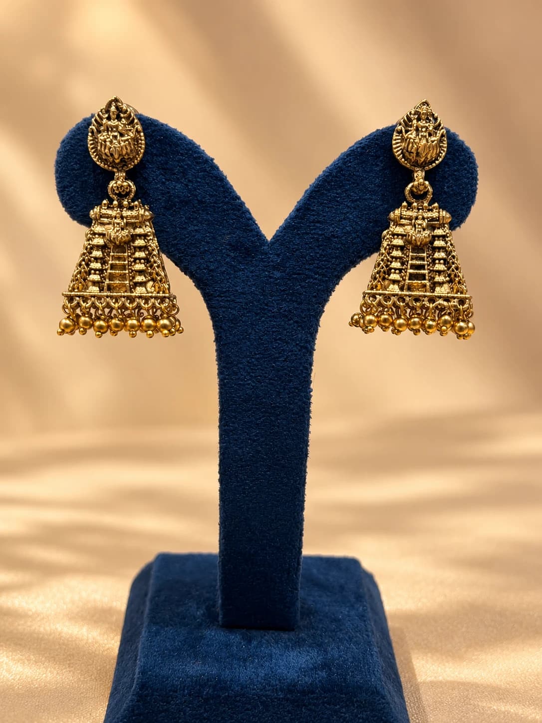 Pair of intricate gold Temple Earrings on a blue display stand. Fancy Jewellery with deity and gopuram design, dangling beads.