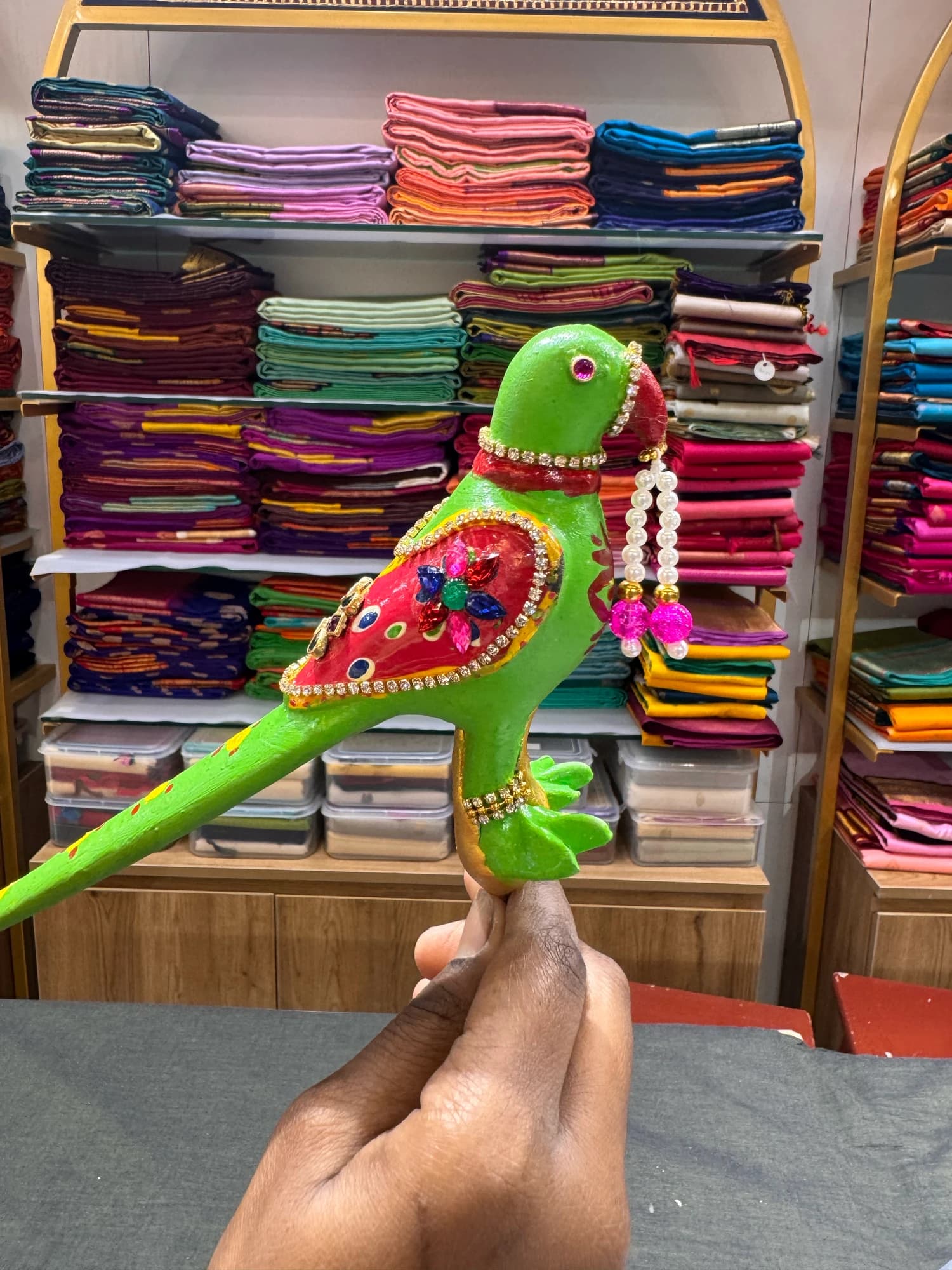 Andal Kili parrot accessory for Bharatanatyam dance, held by hand, against textile shelves.