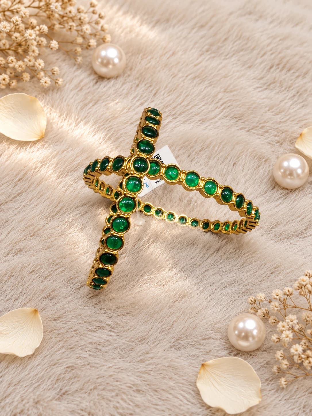 Fancy gold bangles adorned with vibrant green emeralds, displayed on a soft ivory rug with scattered pearls.