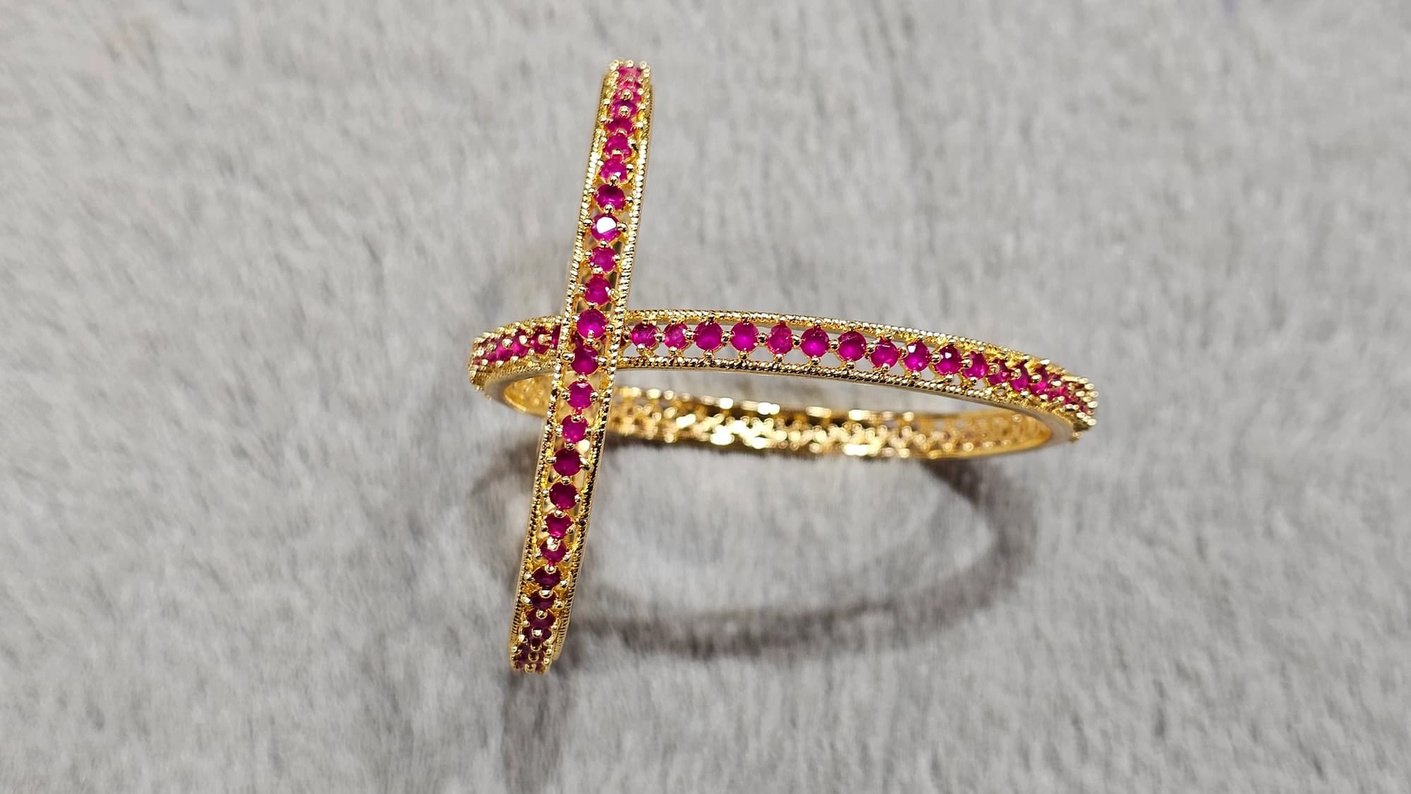 Shanthitailors AD Bangles: Intricate gold-toned bangles with vibrant pink stones, crossed on a soft grey surface.